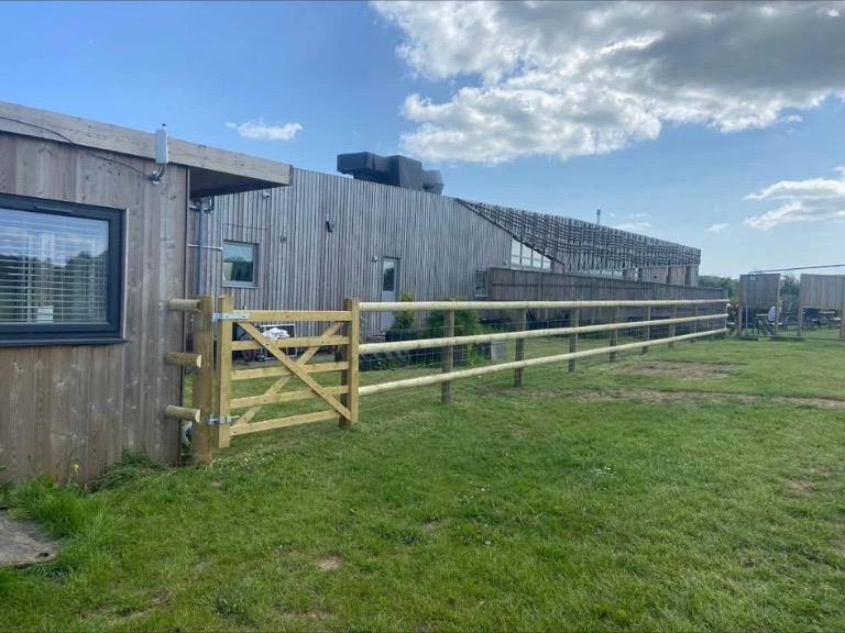 A timber gate and round post and rail fencing beside timber-clad buildings