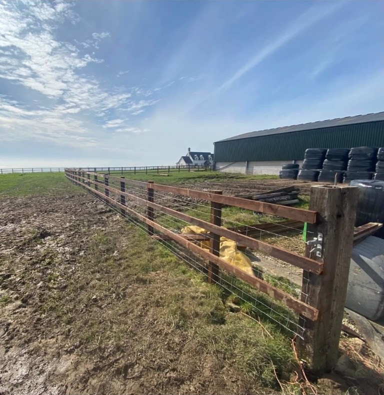 Post and rail fencing with wire mesh alongside a farm building